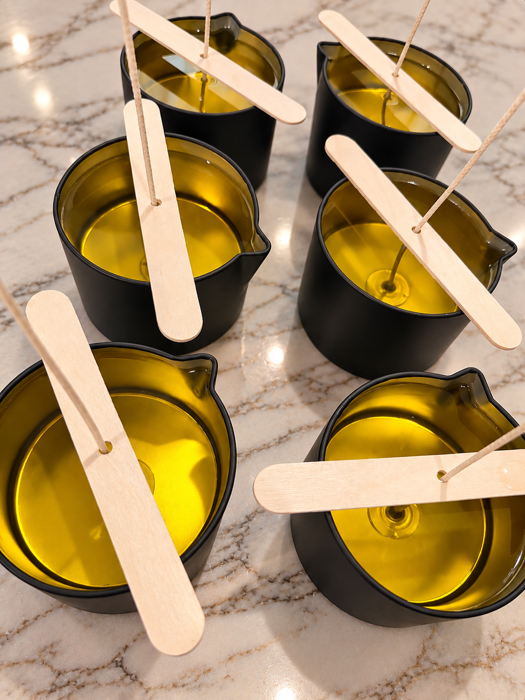 Six freshly poured massage candles in matte black tins with golden oil and centered wicks, resting on a light marble countertop. Made in North Mississippi by Dirty Little Suds