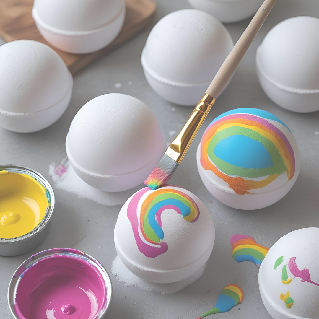 Colorful rainbow designs being hand-painted onto white bath bombs with paintbrushes, paint pots, and unfinished bath bombs in the background — Dirty Little Suds Paint Your Own Bath Bomb Kit.