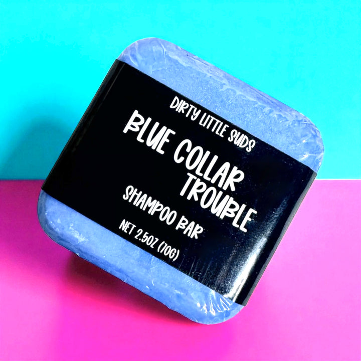 A 2.5 oz Dirty Little Suds Blue Collar Trouble shampoo bar wrapped in a black label, displayed at an angle against a teal and pink background.