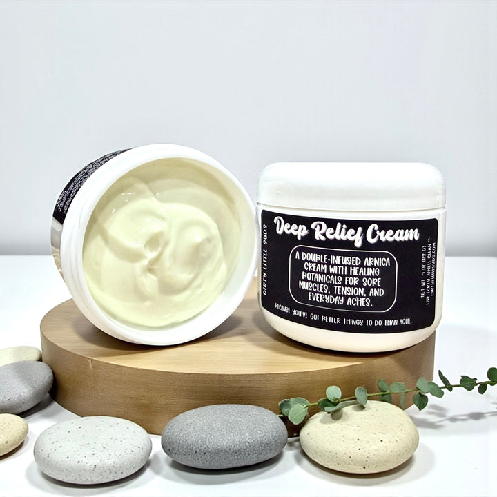 Open jar of Dirty Little Suds Deep Relief Cream showing creamy texture next to sealed jar and spa stones.