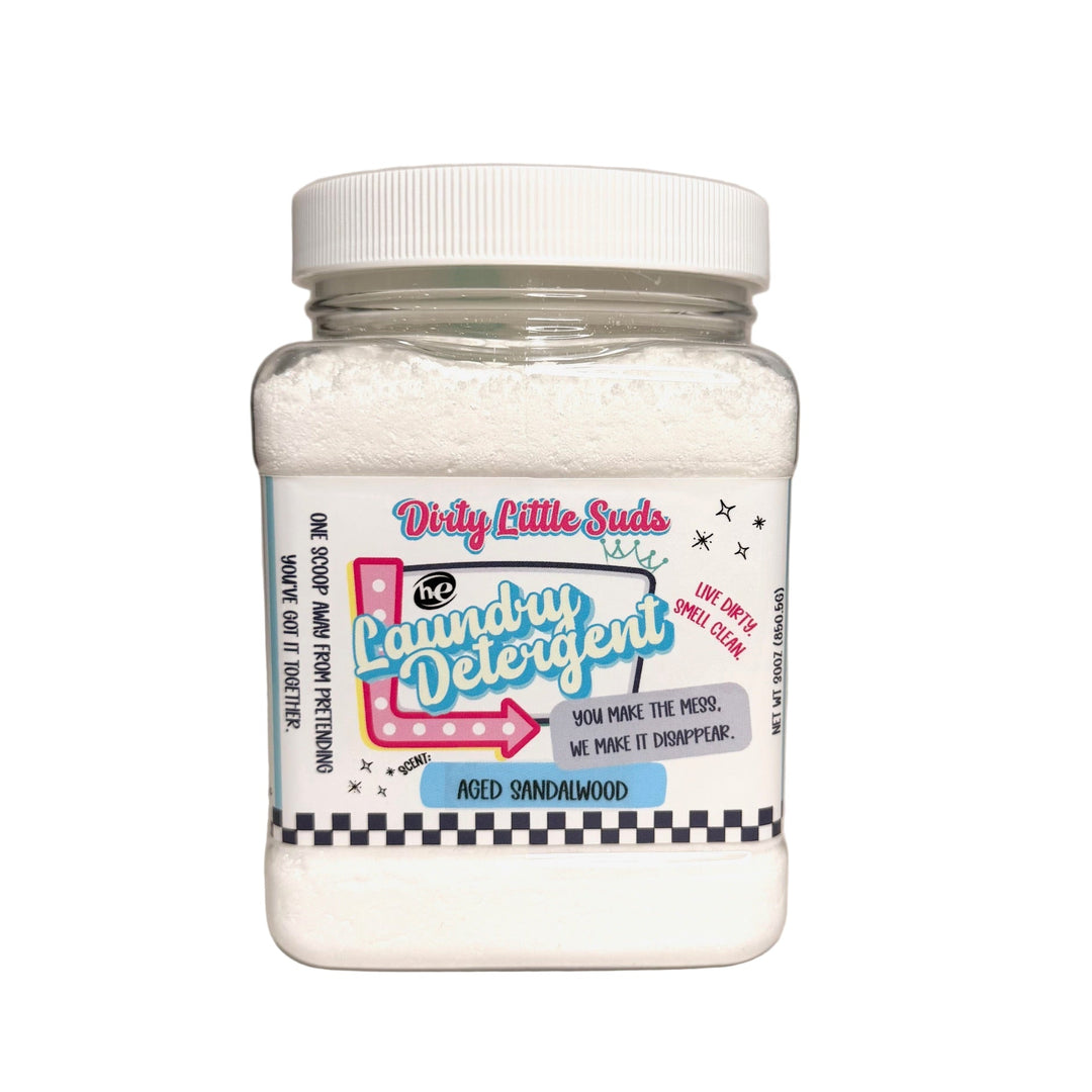 Square clear plastic jar of Dirty Little Suds HE Laundry Detergent in the Aged Sandalwood scent. The detergent is a fine white powder with a bold retro pin-up inspired label featuring turquoise, pink, and black accents. Label tagline reads “You make the mess. We make it disappear.” with checkered trim and playful graphics.