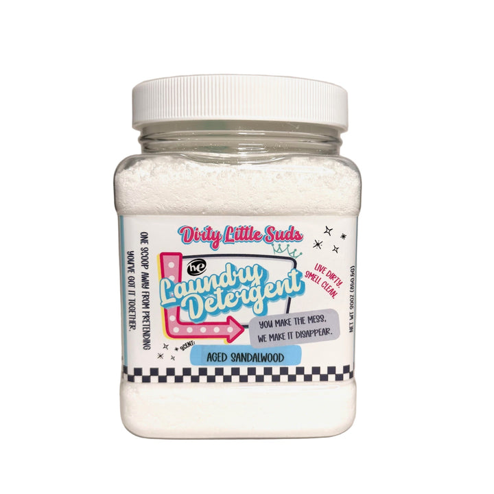 Square clear plastic jar of Dirty Little Suds HE Laundry Detergent in the Aged Sandalwood scent. The detergent is a fine white powder with a bold retro pin-up inspired label featuring turquoise, pink, and black accents. Label tagline reads “You make the mess. We make it disappear.” with checkered trim and playful graphics.