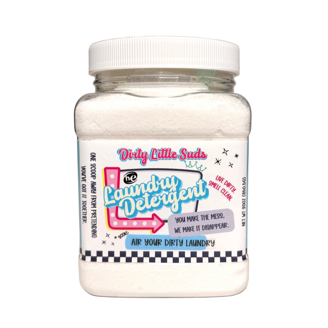 Clear square jar of Dirty Little Suds HE Laundry Detergent in the Air Your Dirty Laundry scent. White powdered detergent with a retro pin-up inspired label in turquoise, pink, and black. A crisp clean fragrance blending fresh linen with cactus bloom for a bold, modern twist on laundry.