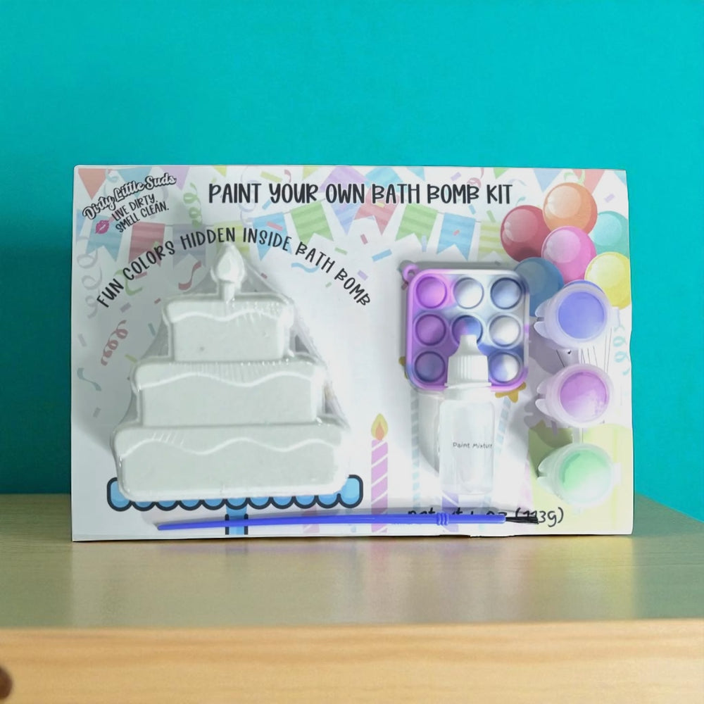 Dirty Little Suds Paint Your Own Bath Bomb Kit – birthday cake bath bomb with hidden colors, paint set, pop-it toy, and brush displayed against teal background.