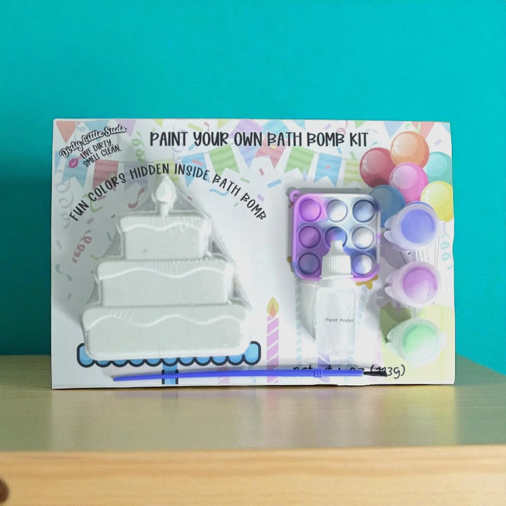 Dirty Little Suds Paint Your Own Bath Bomb Kit – birthday cake bath bomb with hidden colors, paint set, pop-it toy, and brush displayed against teal background.