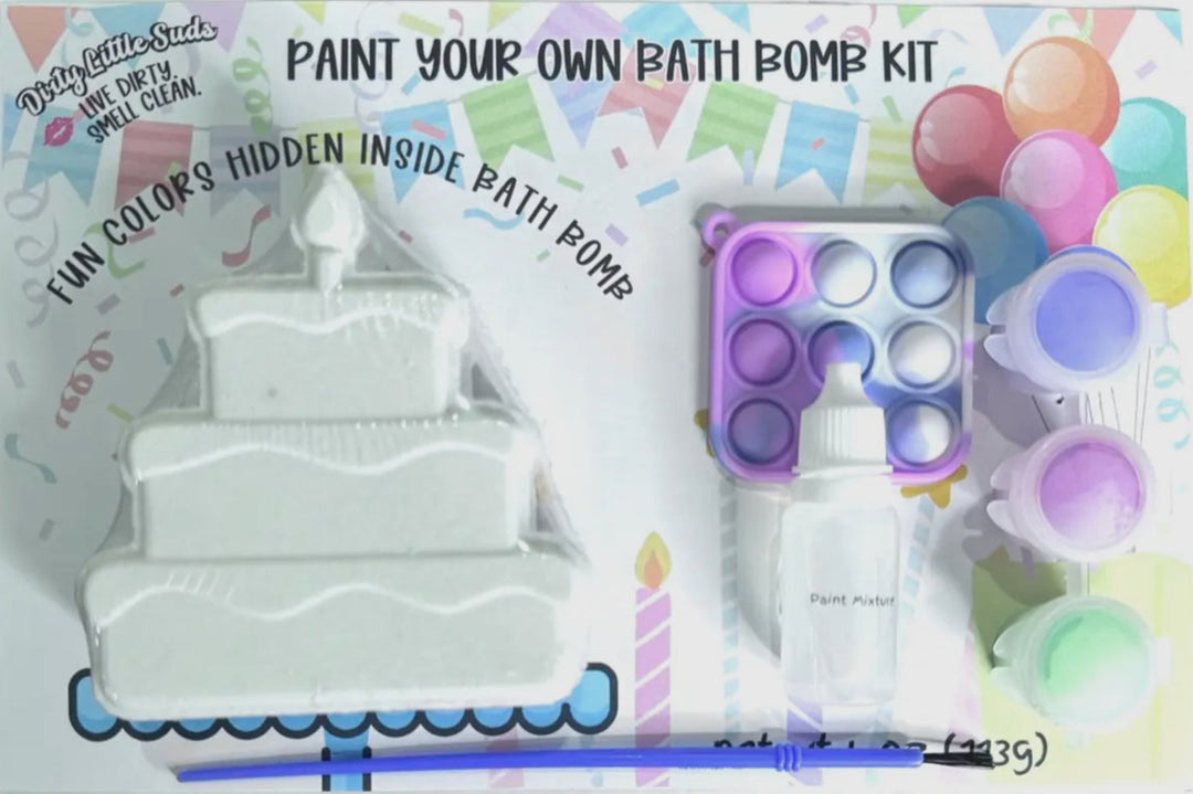 Dirty Little Suds Paint Your Own Bath Bomb Kit featuring a birthday cake bath bomb, colorful paint pots, brush, mixing dropper, and rainbow pop-it fidget toy on party-themed packaging with balloons and confetti.
