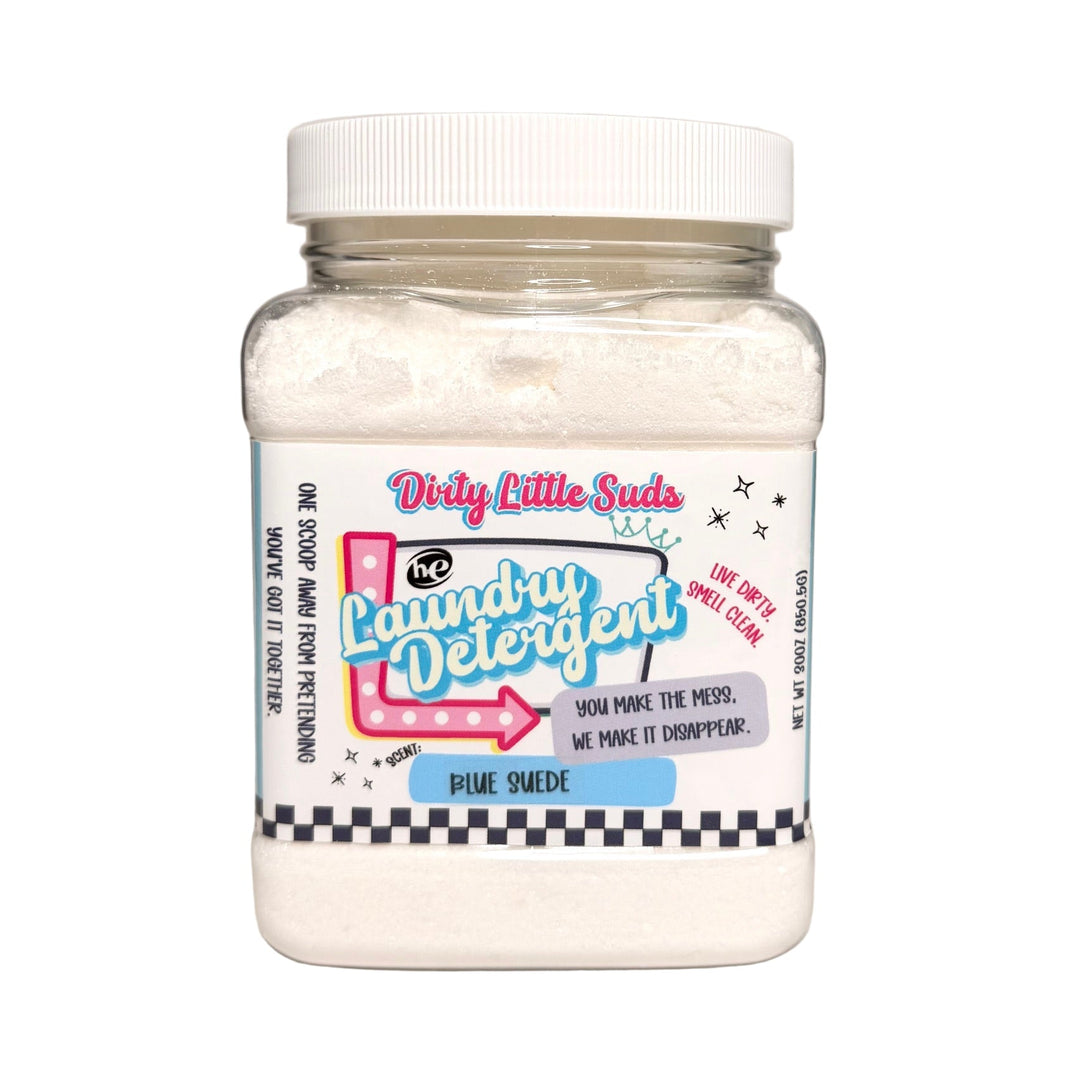 Square clear jar of Dirty Little Suds HE Laundry Detergent in the Blue Suede scent. White powdered detergent with a retro pin-up inspired label in turquoise, pink, and black. A bold fragrance with plum, florals, ocean air, vanilla, and amber for a rich and unforgettable clean.