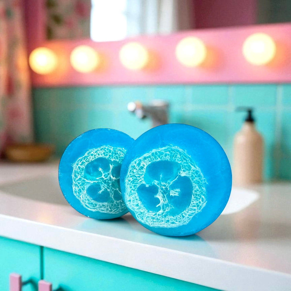 Vibrant blue handmade loofah soap by Dirty Little Suds, scented with blueberry and grape. Exfoliating bar with natural loofah inside, displayed on a retro teal and pink bathroom counter.