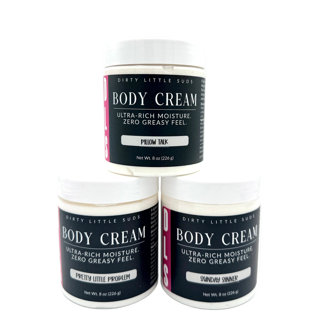 Three 8 oz Dirty Little Suds Body Cream jars featuring the new label design in Pillow Talk, Pretty Little Problem, and Sunday Sinner scents on a white background. Ultra-rich body cream with a non-greasy feel.
