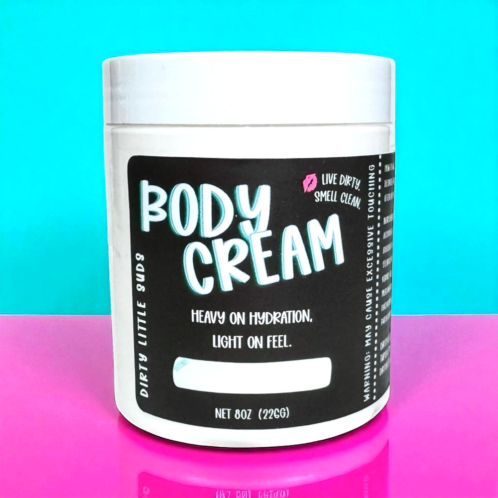Dirty Little Suds Body Cream in an 8oz jar with black label, shown against a turquoise and pink background. Ultra-hydrating, fast-absorbing, non-greasy moisturizer for soft, smooth skin.