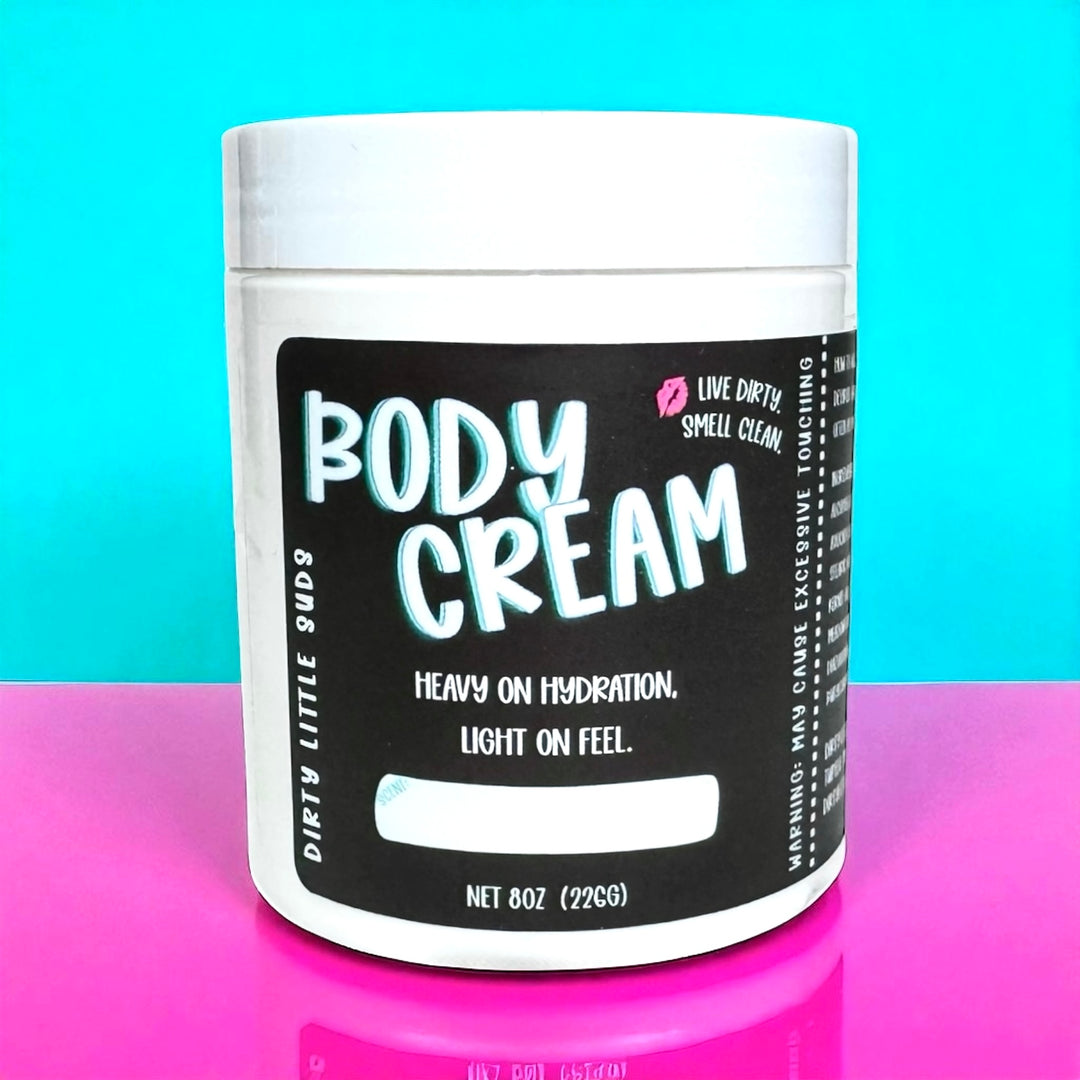 Dirty Little Suds Body Cream in an 8oz jar with black label, shown against a turquoise and pink background. Ultra-hydrating, fast-absorbing, non-greasy moisturizer for soft, smooth skin.