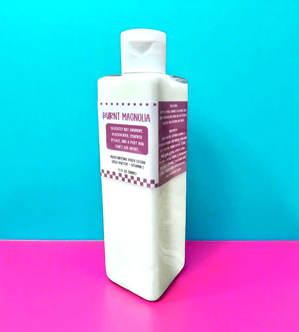 White rectangular 8oz bottle of Dirty Little Suds Burnt Magnolia Body Lotion with a deep pink retro label. The design features playful typography and checkerboard accents, describing the scent as bold and unconventional with notes of blackberry, charred petals, and mystery. Displayed against a turquoise and hot pink background for a vintage pin-up vibe.