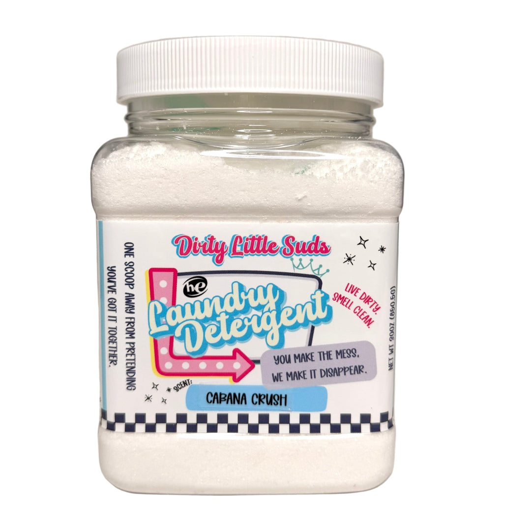 Clear square jar of Dirty Little Suds HE Laundry Detergent in the Cabana Crush scent. White powdered detergent with a retro pin-up inspired label in hot pink, turquoise, and black. A fizzy tropical blend of pineapple, banana, champagne, and sweet fruits that makes laundry smell like vacation.