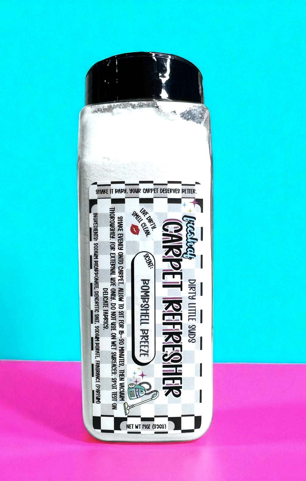 Tall rectangular shaker bottle of Dirty Little Suds Carpet Refresher with a black cap, filled with white powder. The retro label features bold typography and checkered accents in black, turquoise, and purple, reading “Shake it baby, your carpet deserves better.” The product scent is Bombshell Breeze. The background is bright turquoise and hot pink for a playful, vintage pin-up vibe.