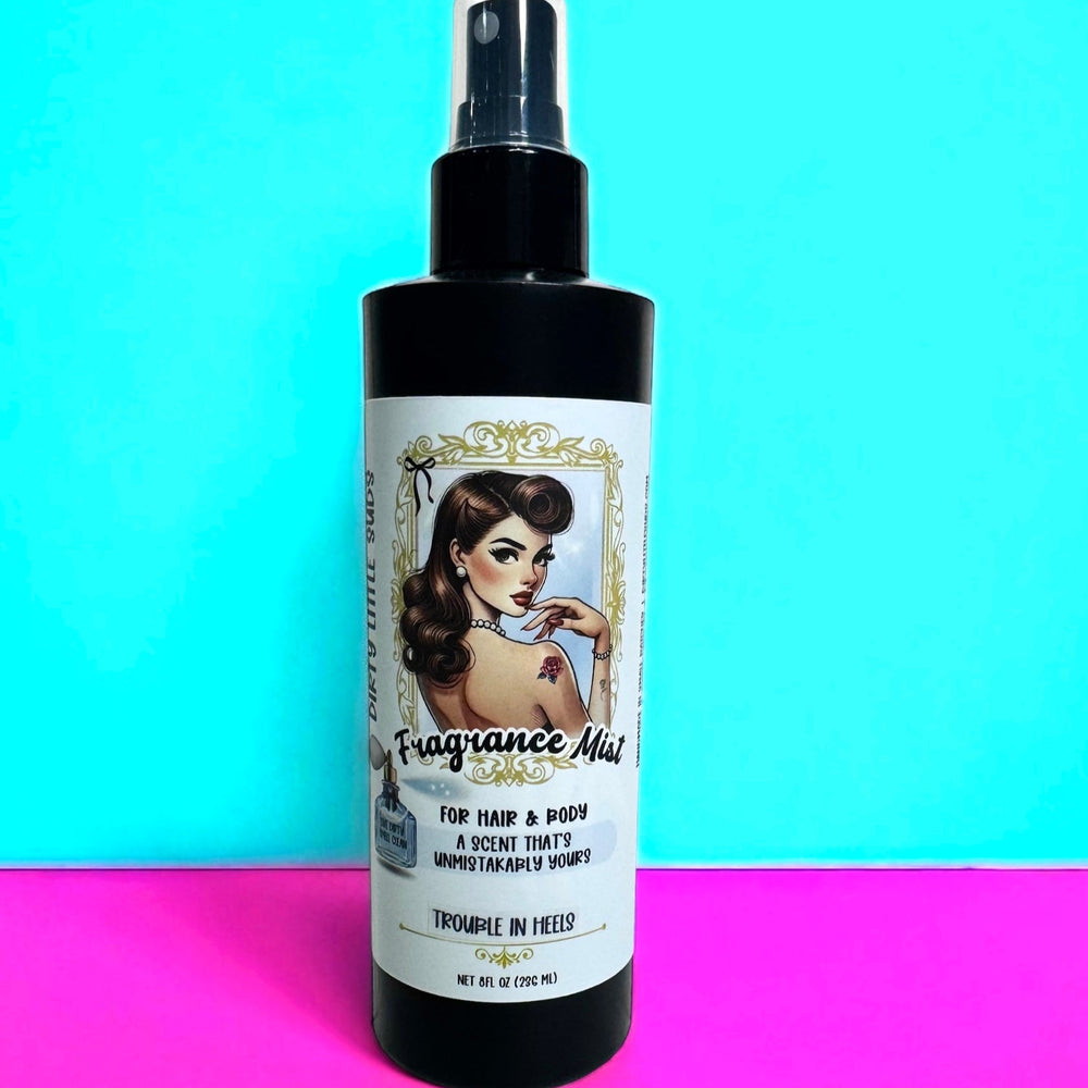 Black 8oz spray bottle of Dirty Little Suds Fragrance Mist with a retro pin-up style label. Designed as a versatile hair and body spray, the bottle is displayed against a turquoise and hot pink background. Perfect for adding a long-lasting, refreshing scent to skin and hair, available in multiple fragrance options.