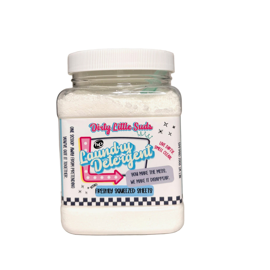 Clear square jar of Dirty Little Suds HE Laundry Detergent in the Freshly Squeezed Sheets scent. Fine white powdered detergent with a retro pin-up inspired label in pink, turquoise, and black. A crisp, airy floral and musk fragrance that smells like freshly washed linen and clean laundry.