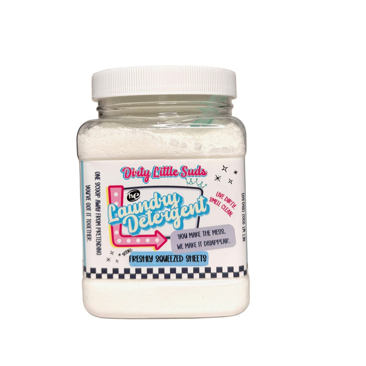 Clear square jar of Dirty Little Suds HE Laundry Detergent in the Freshly Squeezed Sheets scent. Fine white powdered detergent with a retro pin-up inspired label in pink, turquoise, and black. A crisp, airy floral and musk fragrance that smells like freshly washed linen and clean laundry.