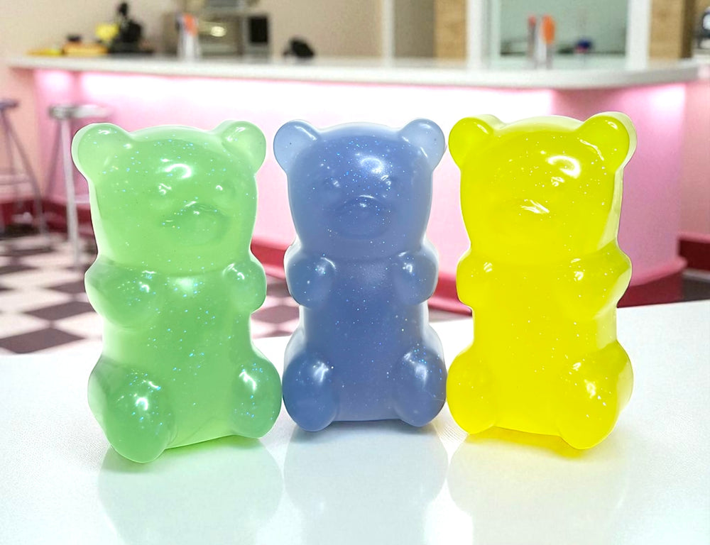 Trio of glitter gummy bear soaps by Dirty Little Suds in green, purple, and yellow, displayed on a retro diner-style counter. Handmade novelty soaps with biodegradable mica glitter, scented in Smell the Rainbow, Sour Candy, and Fruit Loops.