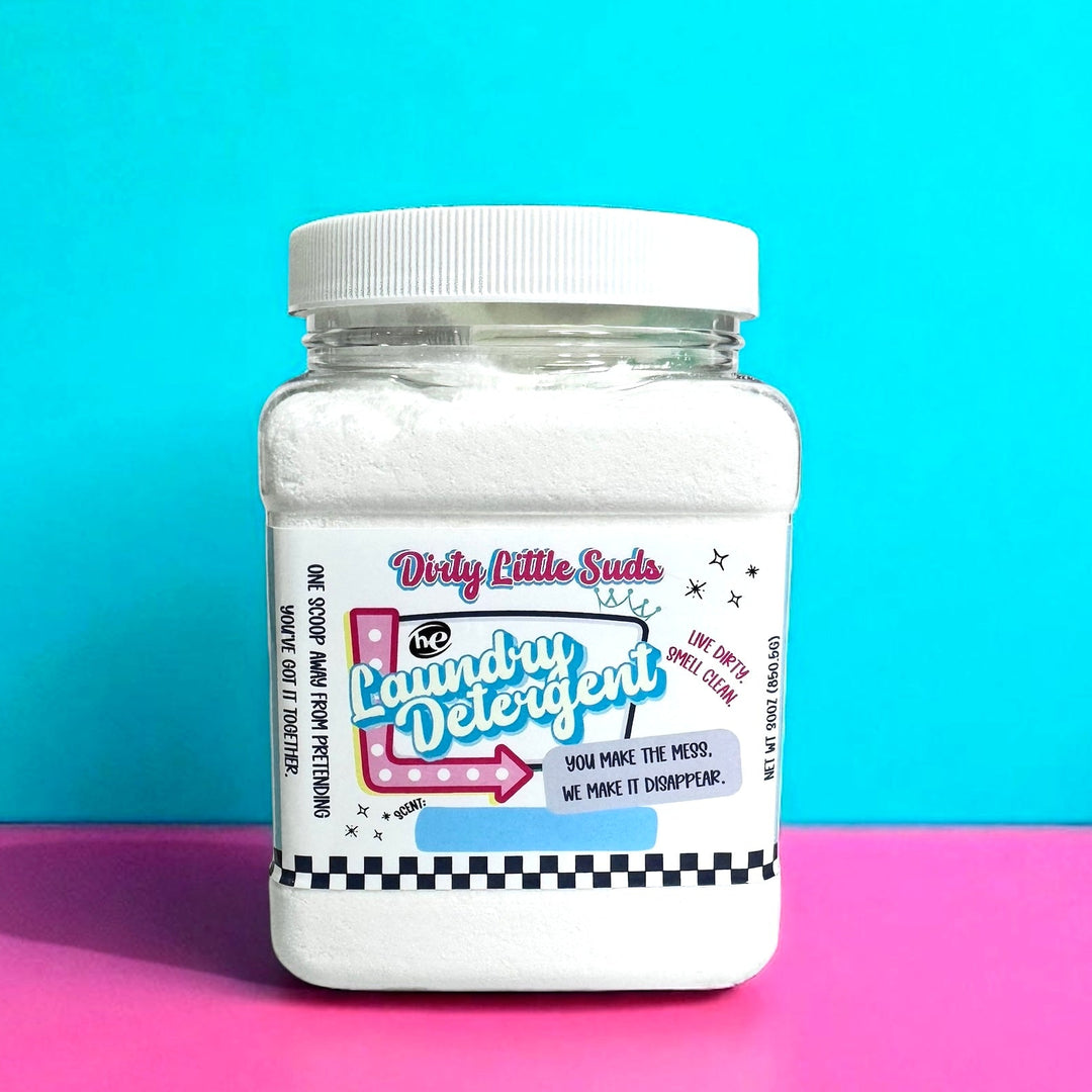 Square plastic jar of Dirty Little Suds HE Laundry Detergent with a white lid, filled with powder detergent. The label features a retro pin-up inspired design in hot pink, turquoise, and black with playful typography. The text reads “You make the mess. We make it disappear.” against a bright two-tone turquoise and pink background.