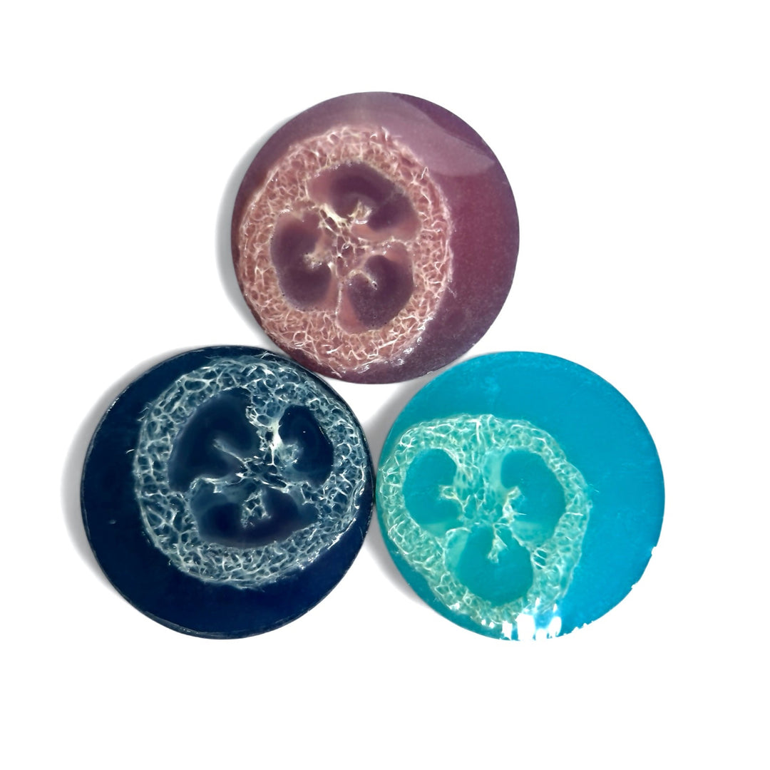 A group shot of three round loofah soaps from Dirty Little Suds in bold jewel tones. The cherry soap is scented in Twisted Cherry, the dark navy soap in Black Raspberry Vanilla, and the bright turquoise soap in Blueberry No. 9. Each bar features a natural loofah slice embedded inside for gentle exfoliation.