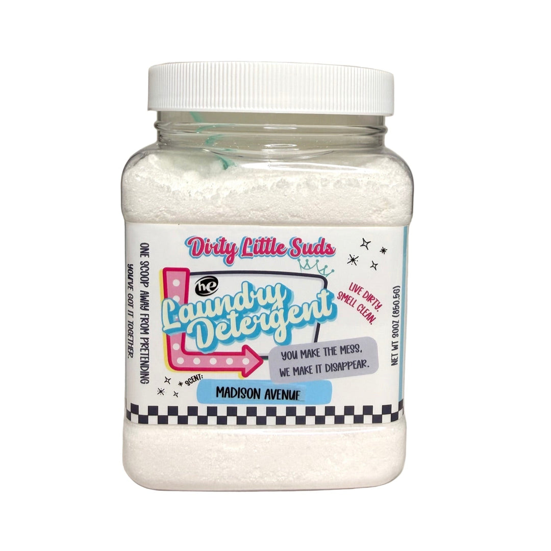 Clear square jar of Dirty Little Suds HE Laundry Detergent in the Madison Avenue scent. Fine white powdered detergent with a retro pin-up inspired label in turquoise, pink, and black. A sophisticated blend of citrus, fig, and cedarwood reminiscent of a luxury Italian hotel.
