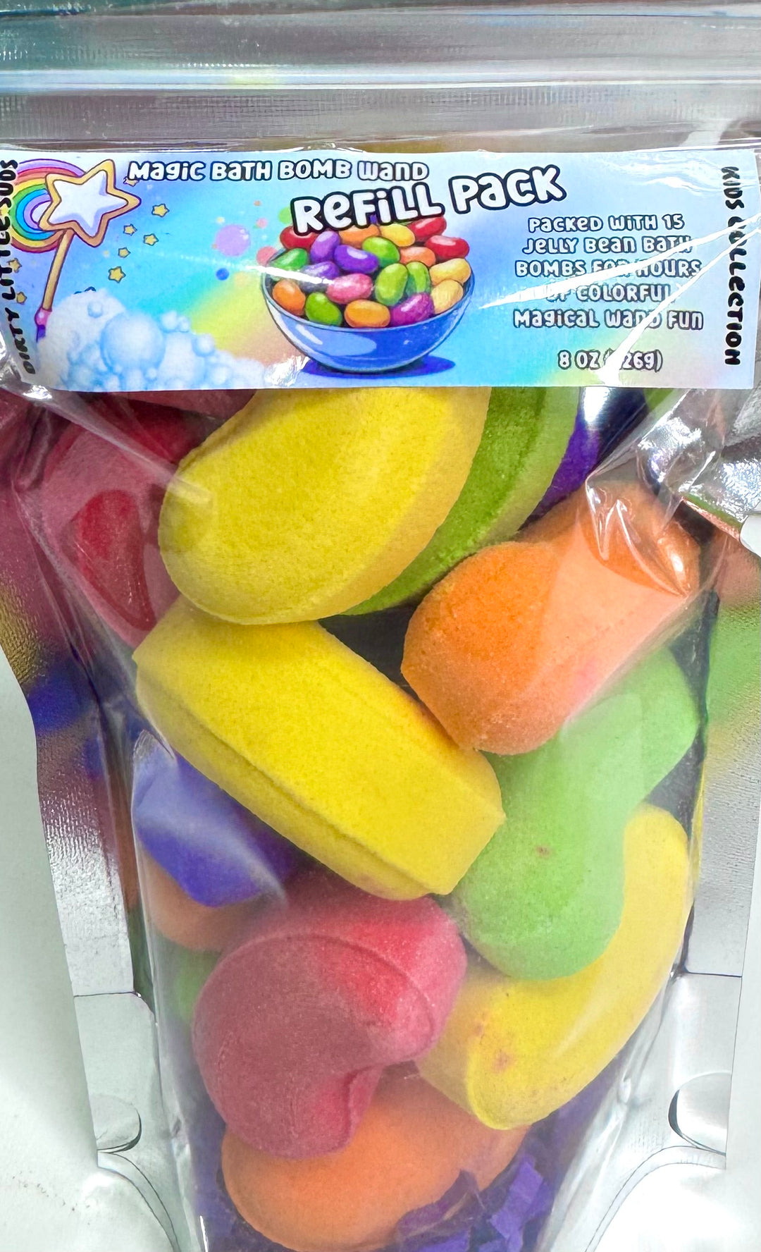 Close-up of the Dirty Little Suds Magic Bath Bomb Wand Refill Pack showing a clear pouch filled with 15 colorful jelly bean–shaped bath bombs in yellow, orange, pink, green, and purple. The playful refill label reads “Magic Bath Bomb Wand Refill Pack” with a rainbow wand and candy bowl graphic, highlighting 8oz of fun bath bomb beans designed for imaginative water play.