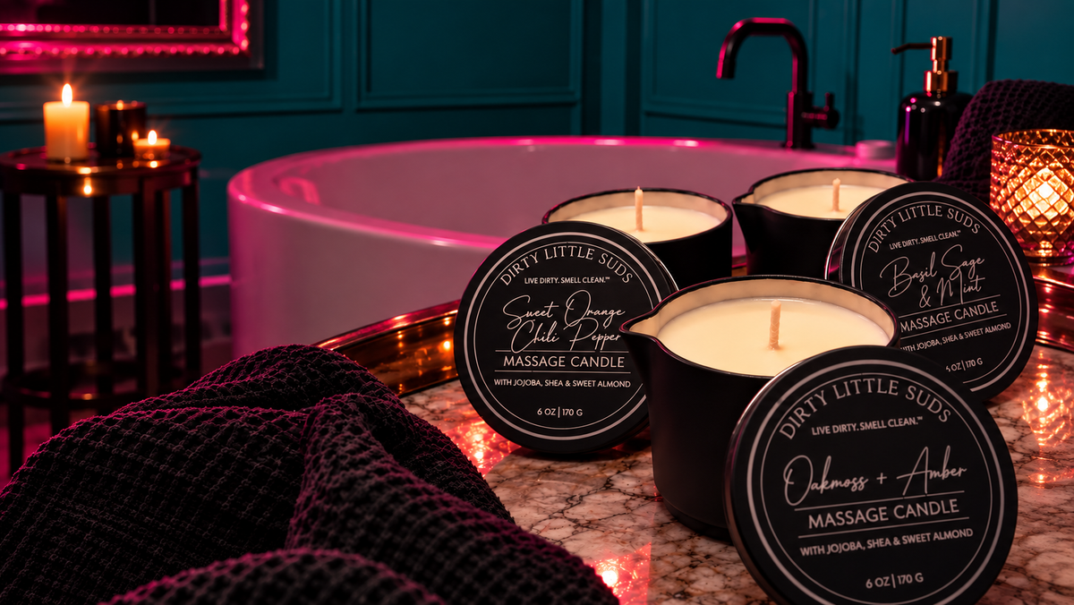 Dirty Little Suds 6 oz massage candles in black pour-spout vessels, featuring Oakmoss + Amber, Basil Sage + Mint, and Sweet Orange + Chili Pepper, displayed beside a bathtub with deep turquoise walls and pink accent lighting