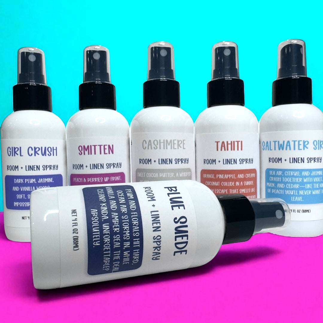 Lineup of Dirty Little Suds Room & Linen Sprays in white bottles with black spray tops, displayed against a turquoise and hot pink background. Scents shown include Girl Crush, Smitten, Cashmere, Tahiti, and Saltwater Siren, with one Blue Suede bottle laying in front. Labels feature bold, playful typography and scent descriptions in colorful blocks.