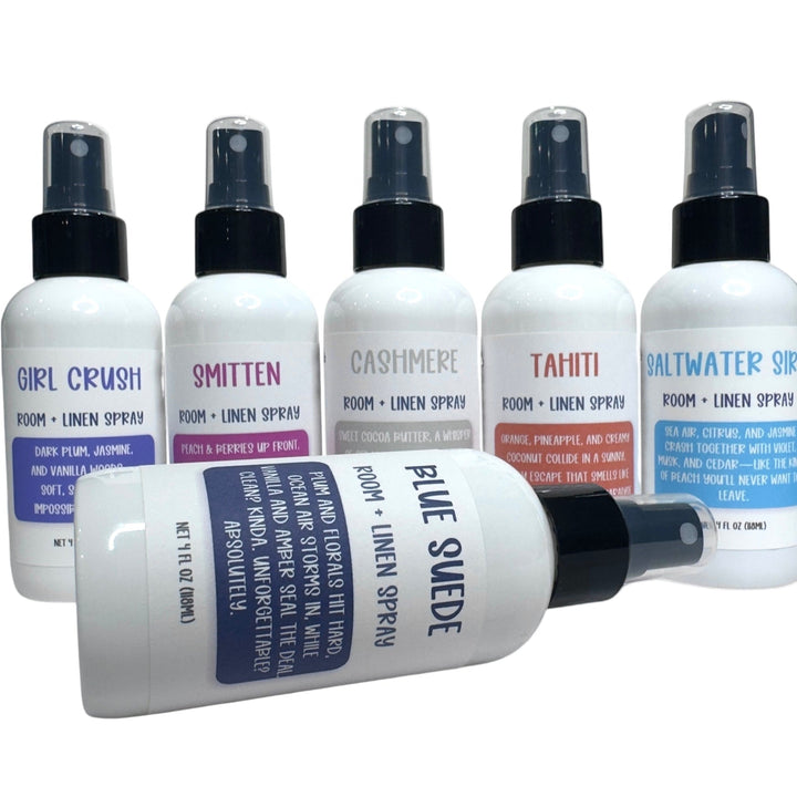 White spray bottles of Dirty Little Suds Room + Linen Spray with bold retro labels in multiple scents including Girl Crush, Smitten, Cashmere, Tahiti, Saltwater Siren, and Blue Suede.