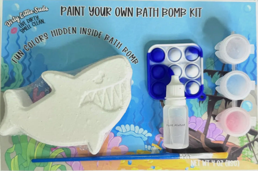 Dirty Little Suds Paint Your Own Bath Bomb Kit featuring a shark bath bomb, paint pots, brush, mixing dropper, and blue pop-it fidget toy on ocean-themed packaging.