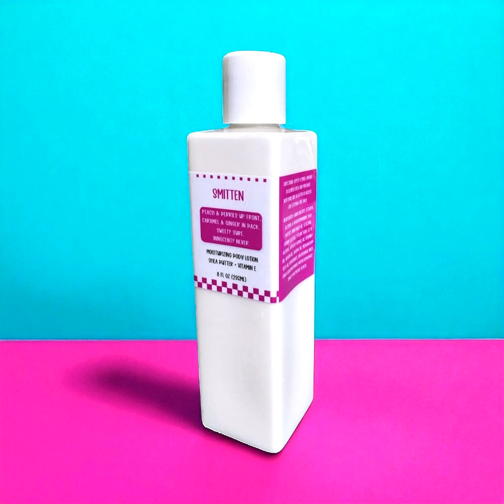 White rectangular bottle of Dirty Little Suds Smitten Body Lotion with a hot pink label featuring cheeky retro typography. The label describes the scent as “Peach & berries up front, caramel & ginger in back. Sweet? Sure. Innocent? Never.” Displayed against a bright turquoise and pink background for a bold, vintage pin-up vibe.