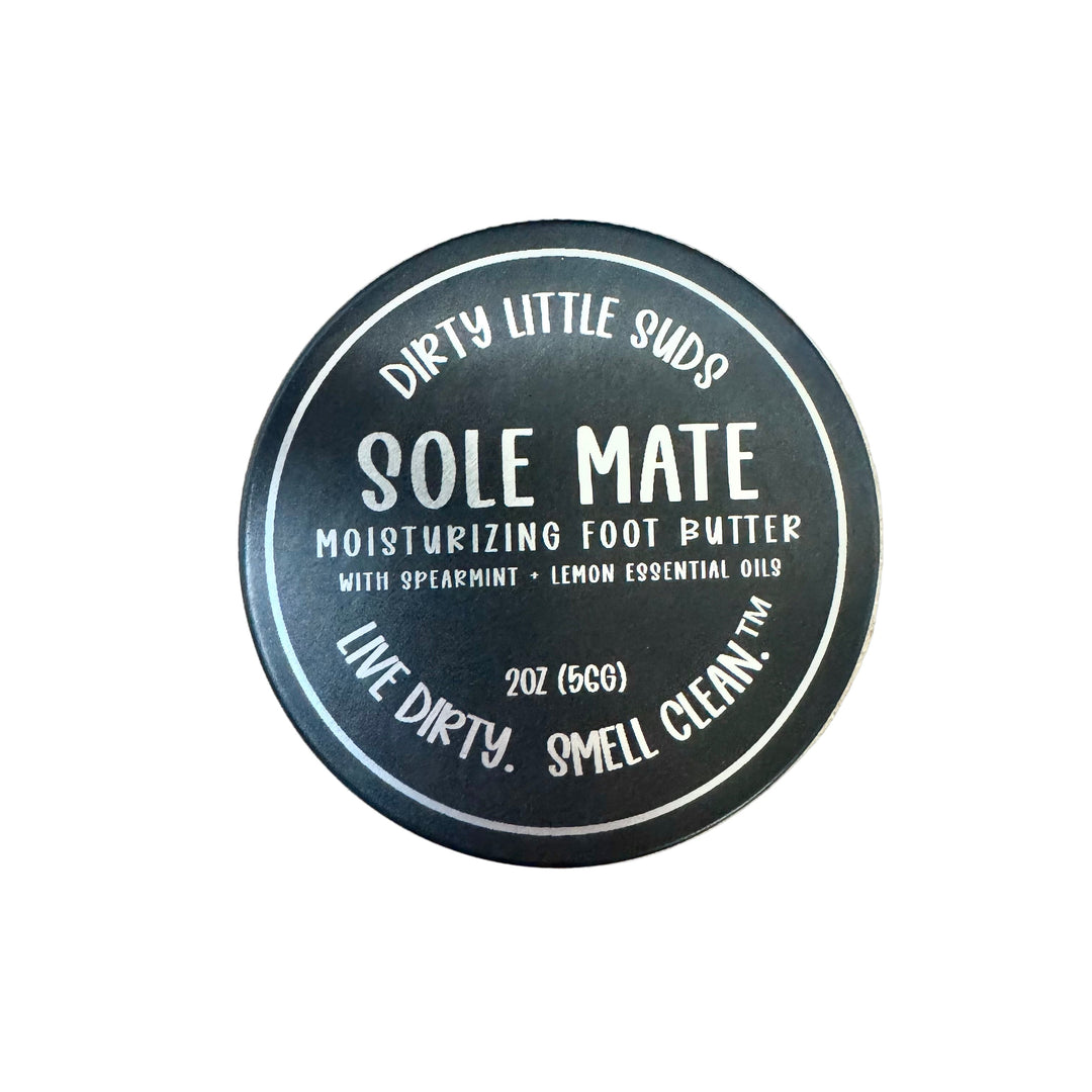 Black engraved tin of Dirty Little Suds Sole Mate Moisturizing Foot Butter, featuring spearmint + lemon essential oils, with retro-style label design shown on lid.
