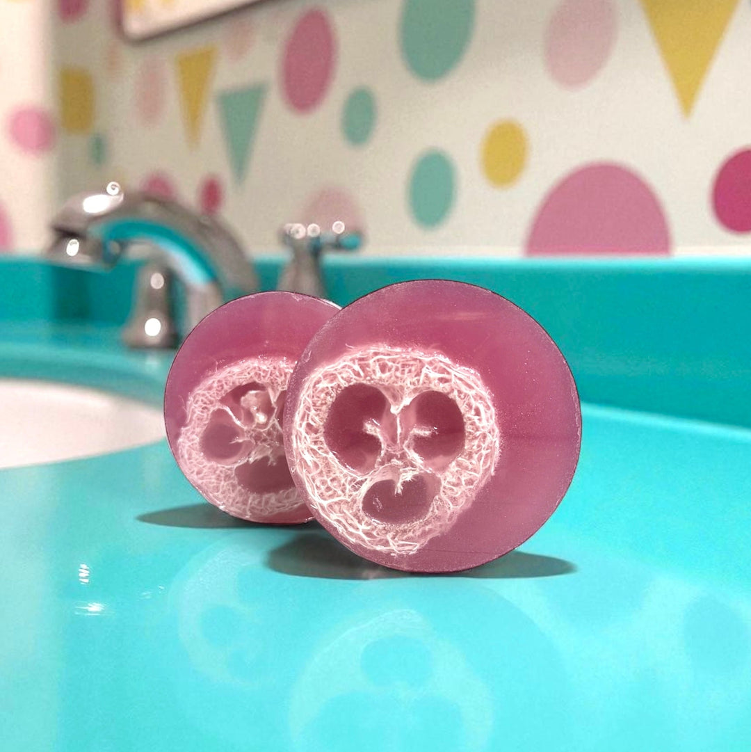 Cherry-red handmade loofah soap by Dirty Little Suds, scented with cherry lemonade. Exfoliating loofah soap bar with natural loofah center, photographed on a retro teal sink with colorful geometric bathroom backdrop.