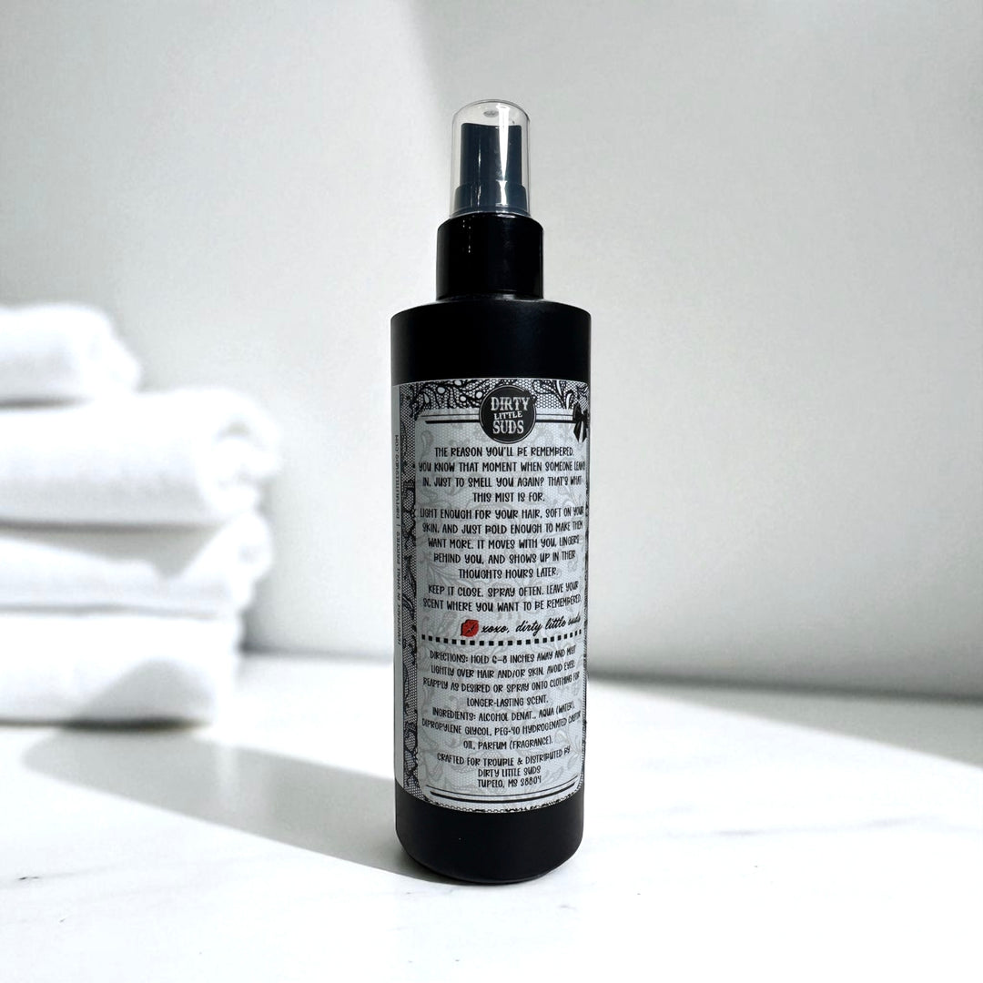Back of a Dirty Little Suds fragrance mist bottle showing detailed label with ingredients, directions for use, and brand info. Retro pin-up inspired design on a sleek black spray bottle, part of the Dirty Little Suds signature body spray collection.