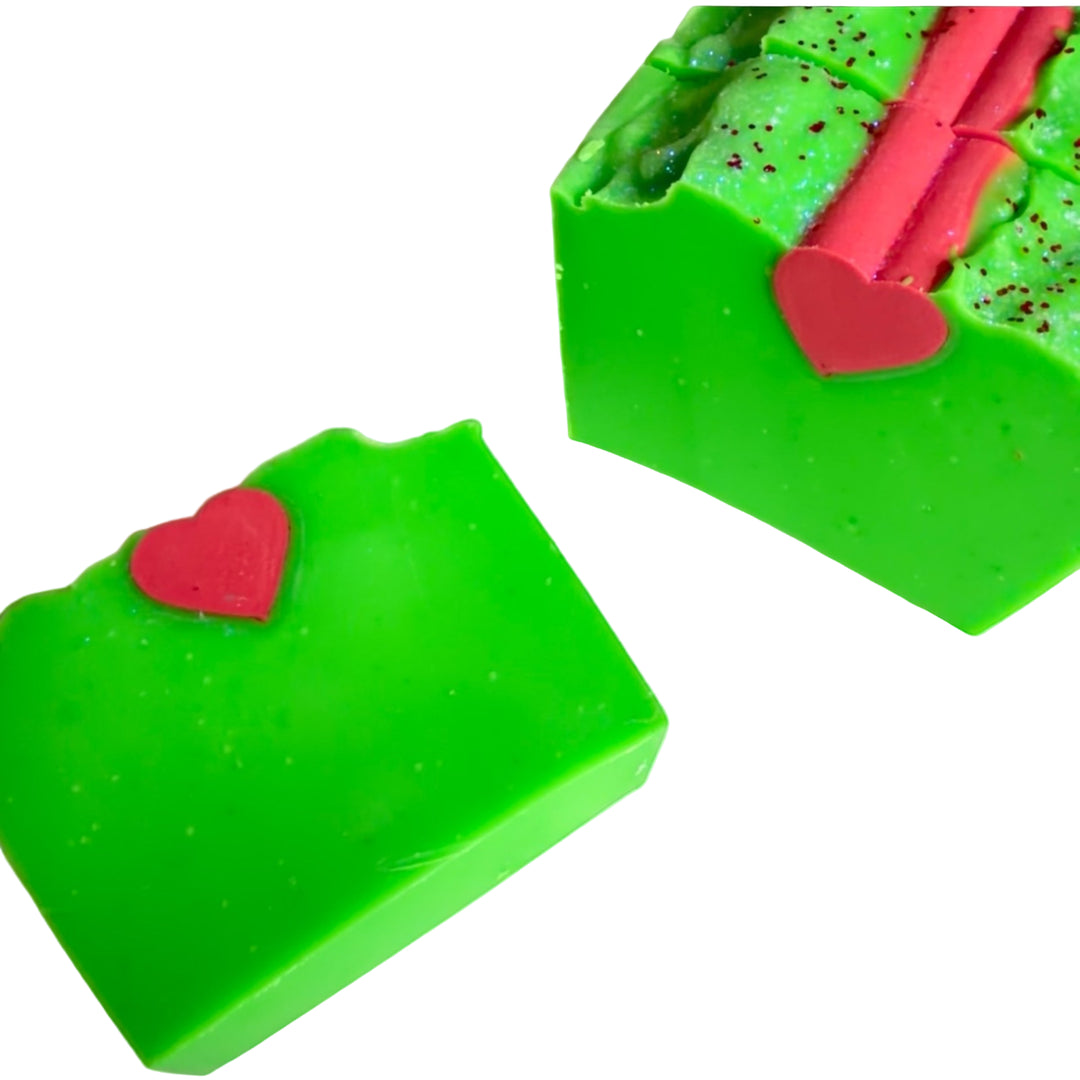 Bright green Extra Grinchy handmade holiday soap bar with a red heart on top, displayed on a festive red background with Christmas tree branches and wrapped gifts. Dirty Little Suds