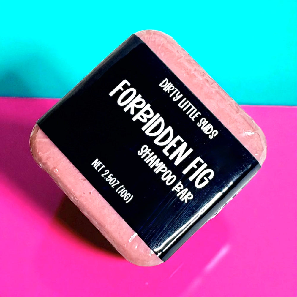 A 2.5 oz Dirty Little Suds Forbidden Fig shampoo bar wrapped in a black label, displayed at an angle against a teal and pink background.