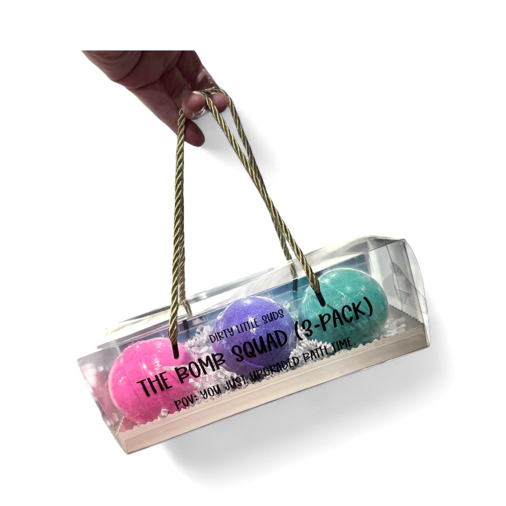 Hand holding a clear gift box with three Dirty Little Suds bath bombs inside. The Bomb Squad 3-Pack features Pressed Petals, Midnight Tea, and Burnt Magnolia bath bombs packed with sweet almond oil and skin-softening ingredients.