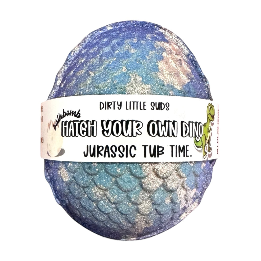 A large shimmering blue and silver dinosaur egg bath bomb wrapped with a Dirty Little Suds label that reads “Hatch Your Own Dino – Jurassic Tub Time.
