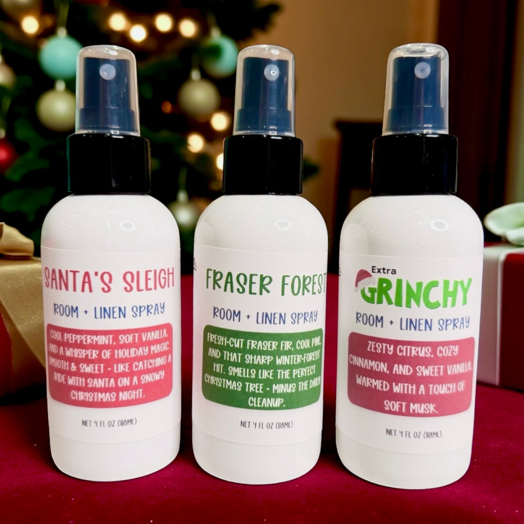 Dirty Little Suds Holiday Room + Linen Sprays – Santa’s Sleigh, Fraser Forest and Extra Grinchy – displayed on a red surface in front of a decorated Christmas tree with lights and ornaments, festive holiday home fragrance scene, Dirty Little Suds