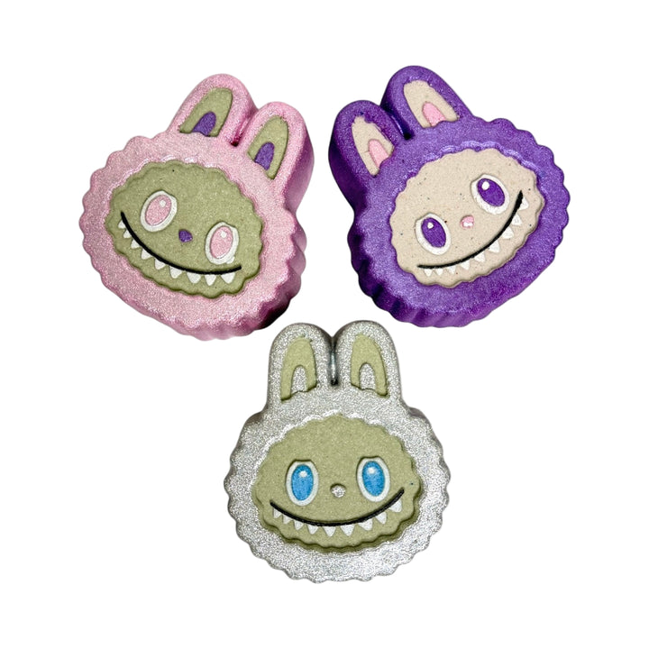 Three hand-painted Labubu Face Bath Bombs in pink, purple, and silver. Each 6oz bomb is scented in Smell the Rainbow and packed with colorful embeds.