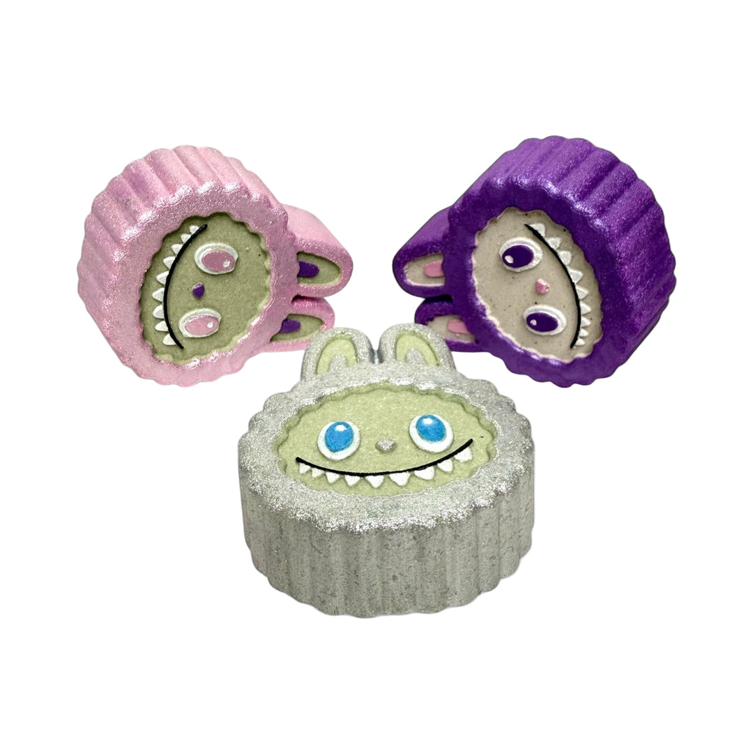 Side view of Labubu Face Bath Bombs in pink, purple, and silver showing their 6oz size and detailed design. Each is scented in Smell the Rainbow with rainbow embeds inside.