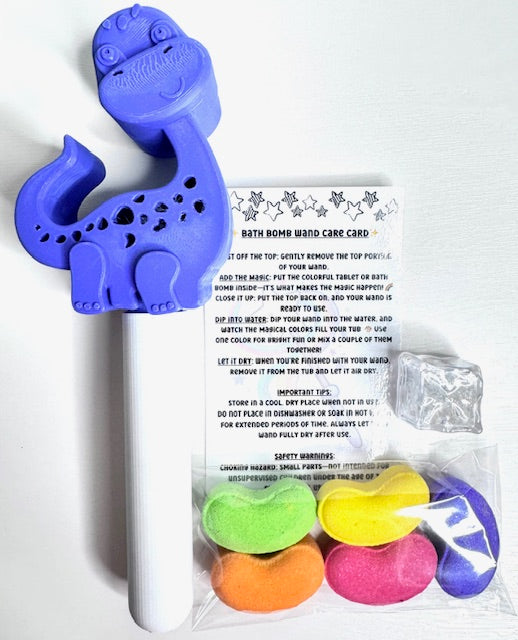 Contents of the Magic Bath Bomb Wand Kit by Dirty Little Suds laid out on a white background. Includes a purple dinosaur bath wand, five rainbow jelly bean bath bombs, a flashing water-activated light cube, and a printed care card with instructions.
