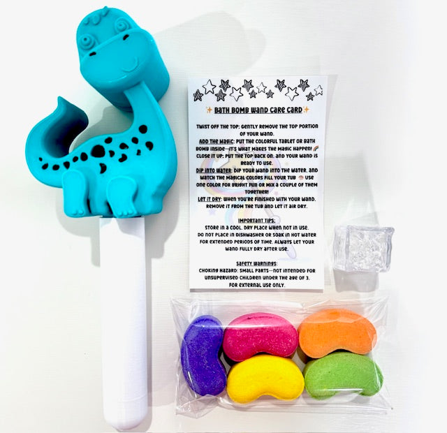 Contents of the Magic Bath Bomb Wand Kit by Dirty Little Suds laid out on a white background. Includes a turquoise dinosaur bath wand, five rainbow jelly bean bath bombs, a flashing water-activated light cube, and a printed care card with instructions.