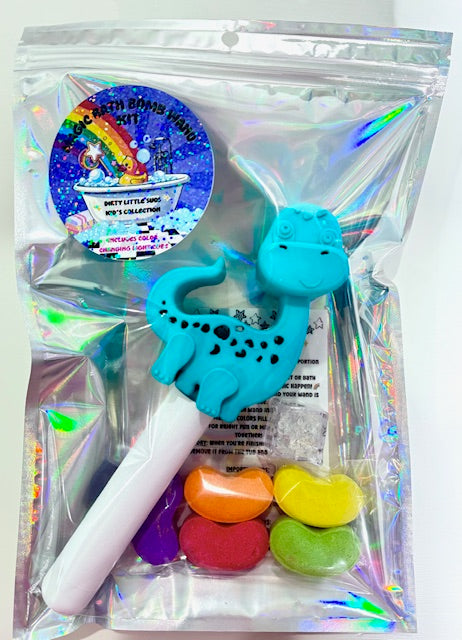 Holographic gift bag containing a Magic Bath Bomb Wand Kit by Dirty Little Suds. The kit includes a blue dinosaur bath wand, colorful jelly bean bath bombs, a flashing light cube, and a holographic rainbow sticker on the front of the bag. Gift-ready bath time set for kids.