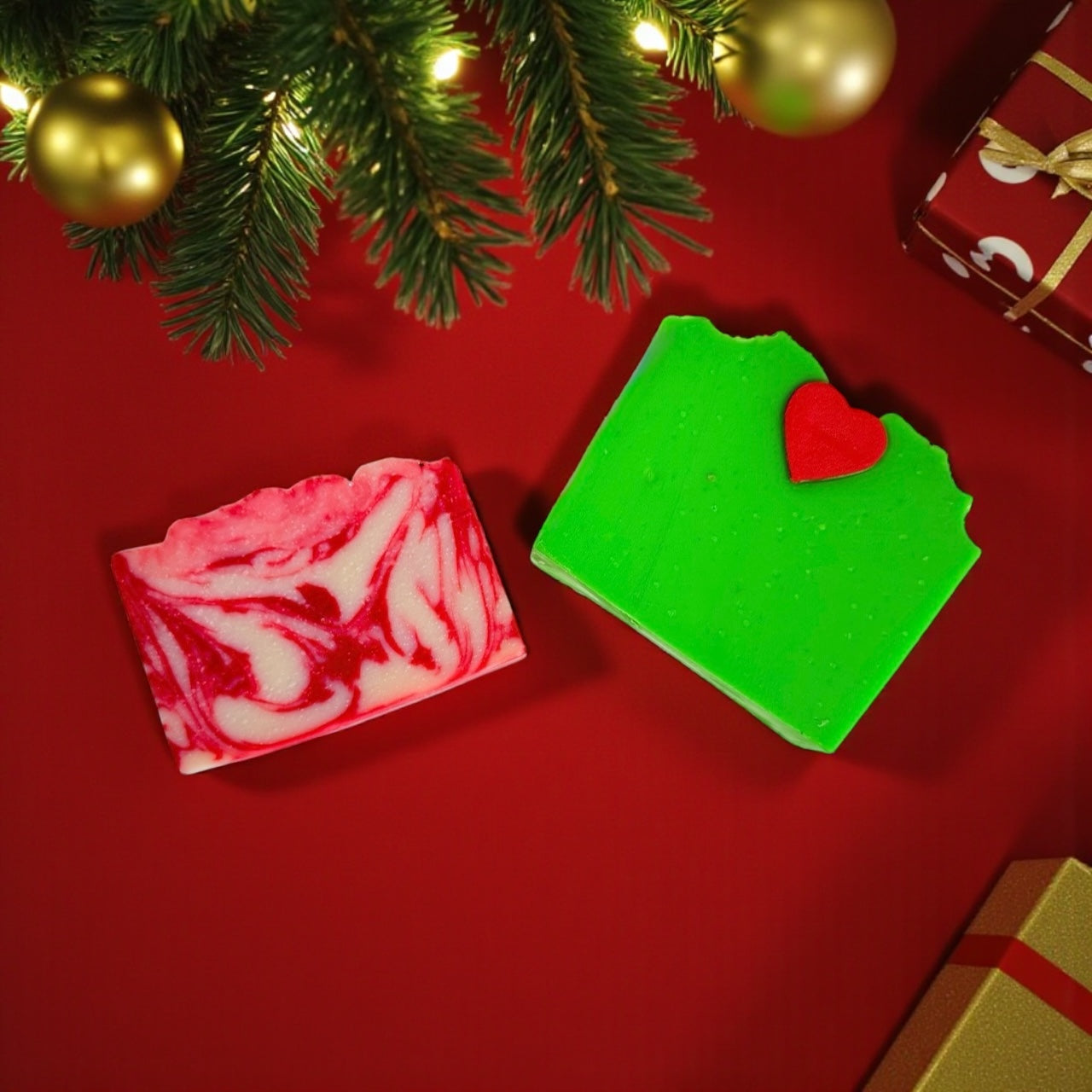 Two handmade holiday soap bars from Dirty Little Suds – a red and white peppermint swirl soap and a bright green Extra Grinchy soap with a red heart on top, displayed on a red background with Christmas tree branches and gifts. Dirty Little Suds