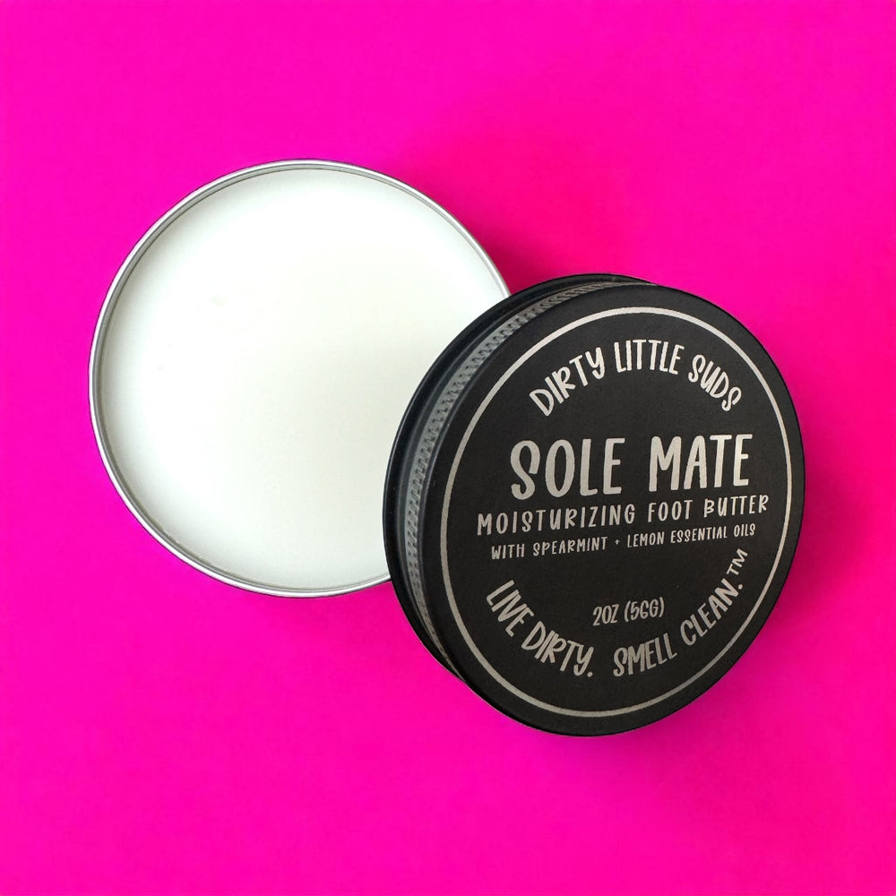 Open engraved tin of Dirty Little Suds Sole Mate Moisturizing Foot Butter with creamy balm inside, black retro lid featuring spearmint + lemon essential oils branding, displayed on bright pink background.