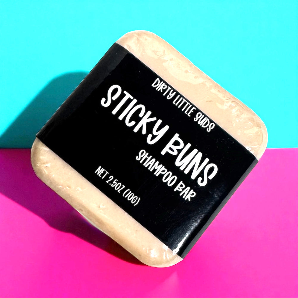 A 2.5 oz Dirty Little Suds Sticky Buns shampoo bar wrapped in a black label, displayed against a bold teal and pink background.