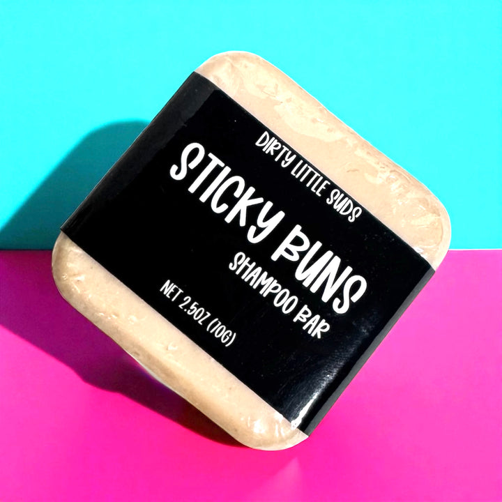 A 2.5 oz Dirty Little Suds Sticky Buns shampoo bar wrapped in a black label, displayed against a bold teal and pink background.