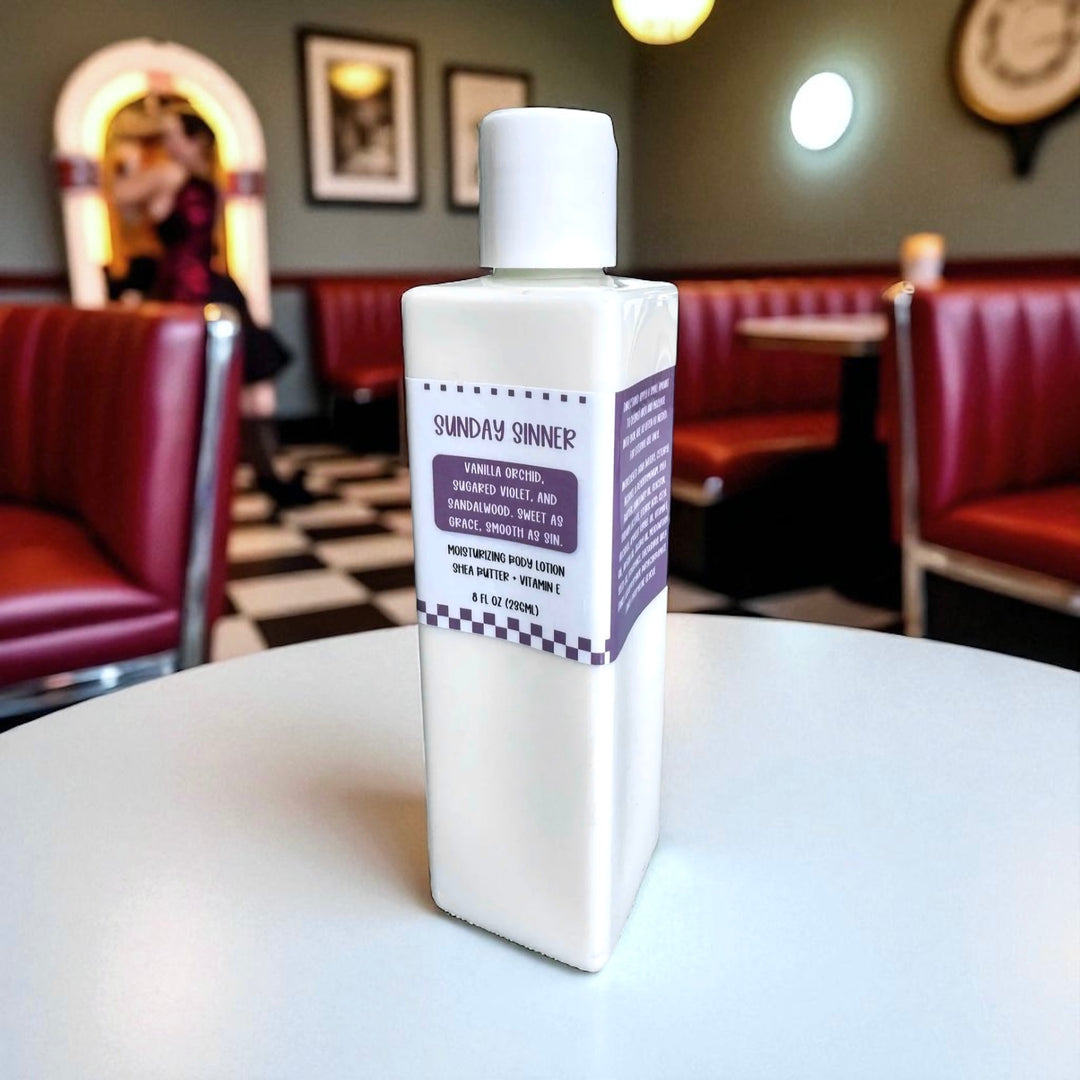 Lifestyle photo of Dirty Little Suds Sunday Sinner Moisturizing Body Lotion on a retro diner table with red booths and vintage décor in the background. Handmade lotion with shea butter and vitamin E, featuring a smooth, warm vanilla and woody gourmand scent.