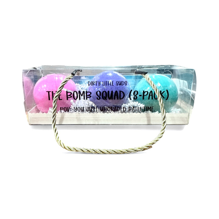 Clear gift box with three colorful handmade bath bombs from Dirty Little Suds. The Bomb Squad 3-Pack includes pink Pressed Petals, purple Burnt Magnolia, and turquoise Midnight Tea bath bombs.